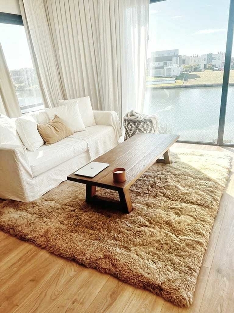 Alfombra de cuero de oveja color avellana sobre un piso de madera, con un sofá blanco y una mesa de madera en un ambiente luminoso junto a una ventana con vista al agua.