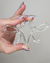 Brinco acrílico transparente em formato de laço, segurado por uma mão com unhas brilhantes.
