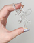 Brinco acrílico transparente em formato de laço sendo segurado por uma mão com unhas decoradas.