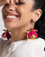 Modelo sorrindo com brinco em metal dourado com design de rosa invertido nas orelhas, destacando detalhes em rosa.