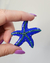 Broche dourado em forma de estrela do mar, esmaltado em azul com strass verde, sendo segurado com uma mão.