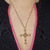 Colar dourado com pingente de cruz religiosa, visível em um fundo com roupa escura e detalhes em rosa.