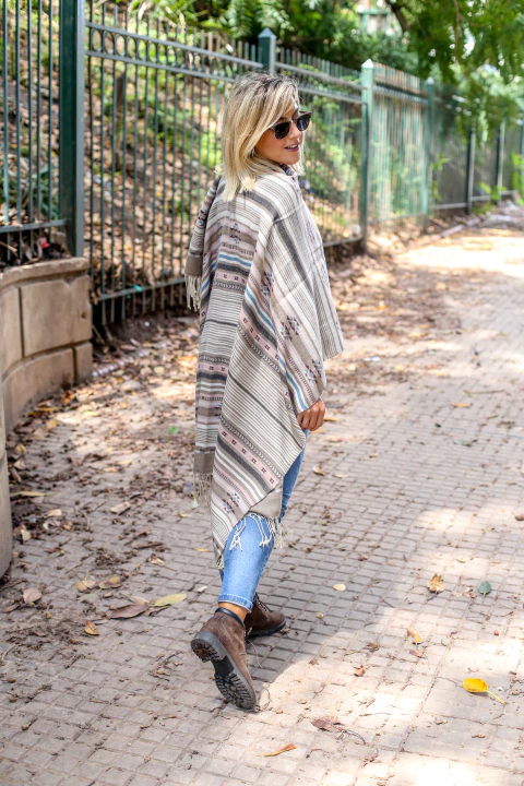 Una mujer caminando por un sendero, vistiendo una ruana de colores neutros y patrones, con jeans y botas. Luce gafas de sol.