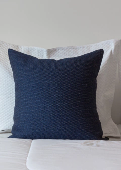 Almofada quadrada de linho tramado na cor azul, posicionada sobre uma cama com lençol branco.