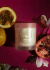 Vela perfumada de magnólia pacífica, cercada por frutas como maracujá e romã, sobre fundo rosa.