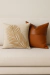 Almofada quadrada bordada com desenho de folha em tom palha ao lado de almofada marrom em couro e tecido, sobre sofá bege claro