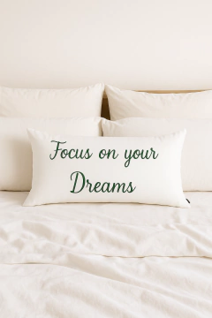 Almofada retangular branca com bordado verde escrito "Focus on your Dreams" sobre cama com lençóis e travesseiros brancos