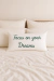 Almofada retangular branca com bordado verde escrito "Focus on your Dreams" sobre cama com roupas de cama brancas