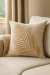Almofada quadrada bege com bordado em forma de folha palha apoiada em sofá claro em ambiente com cortinas neutras
