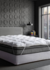 Imagem de um colchão Pillow Top Toque de Pluma com fibras siliconadas, posicionado em um quarto moderno com paredes cinzas e uma janela grande.