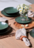 Conjunto de 6 pratos rasos orgânicos na cor verde, decorados com flores brancas em um vaso, sobre uma mesa de madeira com toalha e talheres.