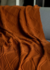 Manta Premium Tricot Cobre em tom laranja, com padrão texturizado, repousada sobre um sofá cinza.