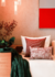 Três almofadas em um sofá com manta verde e flores ao fundo, decorando um ambiente aconchegante.