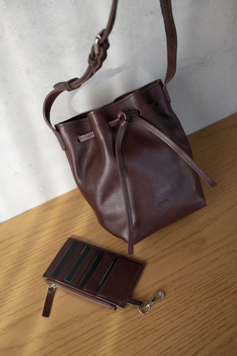 Bandolera de cuero vacuno marrón oscuro estilo marinero con cierre de lazo y cartera organizadora de cuero del mismo color sobre mesa de madera