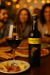 Garrafa de vinho Yellow Tail Shiraz australiano 750ml com taça de vinho tinto e prato com queijos, presunto e palitos, ao fundo pessoas sorrindo em ambiente aconchegante