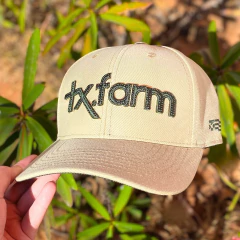 Boné bege com bordado verde escrito tx.farm na parte frontal, segurado por uma mão, com fundo de plantas verdes