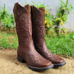 Bota Texana Feminina Goyazes Dallas na cor tabaco, com detalhes em relevo e aplicação de tachas, em fundo de grama verde.