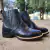 Botas de couro pretas com detalhes em costura e sola de borracha, posicionadas sobre uma superfície de concreto.