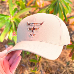 Boné bege Texas Farm com logo camuflado sendo segurado por uma mão, ao ar livre com plantas ao fundo