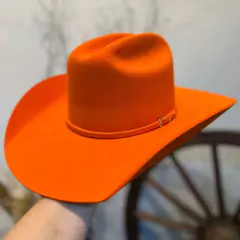 Chapéu Sibu estilo cowboy na cor laranja, com aba larga e detalhe de fita. Fundo desfocado com roda de carroça ao fundo.
