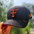 Boné preto com detalhes em laranja neon da marca Texas Farm, modelo Tf672, sendo segurado por uma mão.