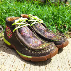 Detalhe dos tênis Goyazes Destroye Café Verde, com design colorido e cadarços verdes, sobre uma superfície de concreto e grama ao fundo.