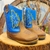 Bota Country Texana Baby Centopeia Azul com detalhes em azul e costura amarela, em um fundo rústico.