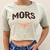 Camiseta cropped feminina bege com estampa "MORS" e detalhe de chifre na parte inferior.