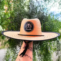 Chapéu de palha feminino da marca Tex Team, com aba larga e detalhe em preto, segurado por uma mão com tatuagens, em meio a folhagens verdes.