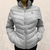 Jaqueta feminina Tuff Puffer cinza com capuz, modelo acolchoado e zíper frontal.