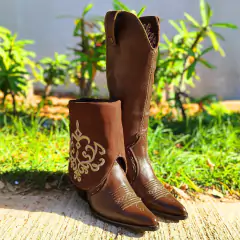 Bota feminina West Country bico fino na cor café, com detalhes bordados, posicionada sobre uma superfície de concreto com grama ao fundo.