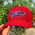Boné vermelho da marca Texas Farm com bordado azul e detalhe na aba, segurado por uma mão.