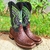 Bota Texana Masculina Cp Boots Exótica na cor marrom, com detalhes em verde e design estilizado, posicionada sobre uma superfície de terra e grama.