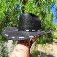 Chapéu infantil boiadeira na cor preta, com detalhes em costura branca, sendo segurado por uma mão em meio a uma vegetação verde.