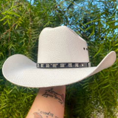 Chapéu country branco de juta com aba larga e faixa preta decorativa segurado por mão com tatuagens, fundo de folhas verdes desfocado