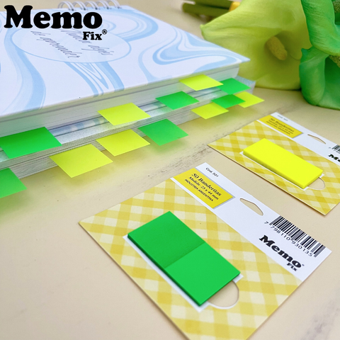 Banderitas autoadhesivas MemoFix verdes y amarillas marcando páginas de un libro, con dos paquetes de banderitas sobre la mesa