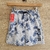 Bermuda con estampado de hojas en tonos azules sobre fondo gris, con cintura elástica y cordón ajustable.