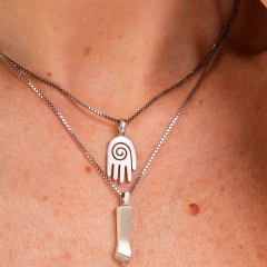Colar de prata com pingentes de mão e figura estilizada, evidenciado em um fundo de pele.