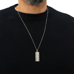 Homem usando um suéter preto com um colar de prata que possui um pendente retangular.