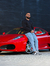 Homem posando ao lado de um carro esportivo vermelho, vestindo uma calça slim fit jeans casual claro e uma camiseta preta.