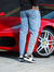 Homem usando calça slim fit jeans casual claro, com tênis pretos e brancos, ao lado de um carro esportivo vermelho.