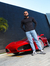 Homem posando ao lado de um carro esportivo vermelho, vestindo calça jeans slim fit claro e camiseta preta.