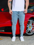 Homem usando calça skinny jeans clara em frente a um carro esportivo vermelho.