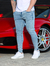 Homem usando calça skinny jeans clara ao lado de um carro esportivo vermelho.