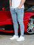 Homem usando calça skinny jeans casual clara, com camiseta branca e tênis brancos, posando ao lado de um carro esportivo vermelho.