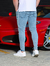 Homem usando calça skinny jeans clara, vista traseira, ao lado de um carro esportivo vermelho.