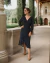 Mulher elegante usando um vestido preto com bolinhas brancas, caminhando em um ambiente externo, cercada por plantas verdes. Ela usa óculos de sol e acessórios delicados.