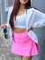 Mujer con cabello largo y rojo, vistiendo un top blanco y una falda corta rosa, sosteniendo gafas de sol.