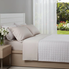 Jogo de lençol king size bege com 4 peças em algodão 100% percal 400 fios, cama arrumada com travesseiros e colcha branca, em quarto claro com vaso de flores ao lado