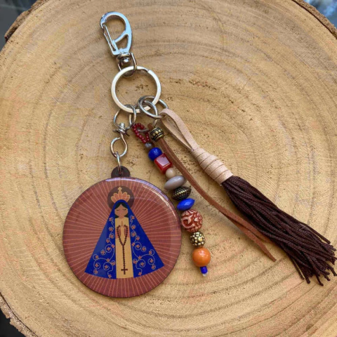 Chaveiro decorativo com imagem estilizada de Nossa Senhora Aparecida, junto com pingentes de miçangas coloridas e tassel marrom, sobre superfície de madeira